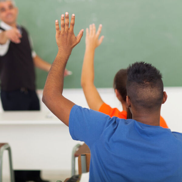 High school students in a classroom raising their hands with the teacher by the chalk board