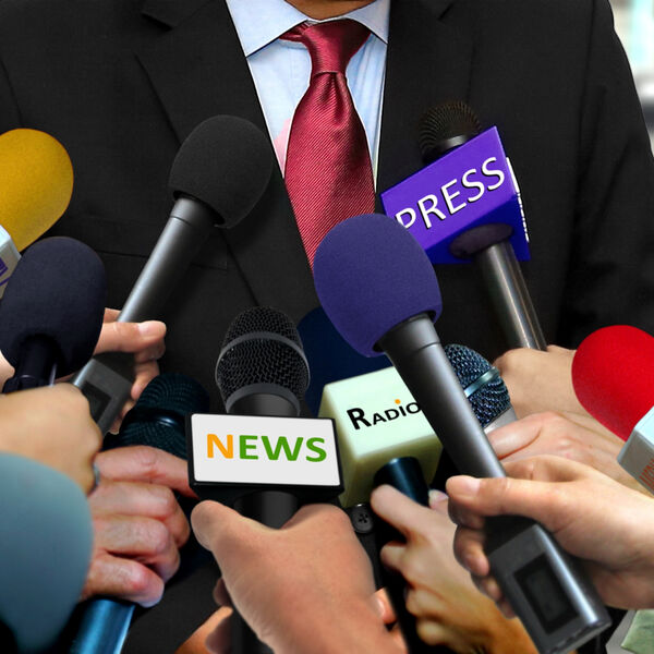 Man in black suit with a red tie surrounded by news reporters