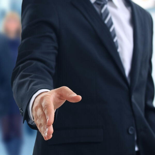 Business man in dark suit holding hand out to shake hands with other business professionals behind him