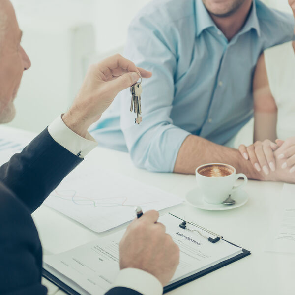 Man in a suit with a pen in one hand over a contract and a set of keys in another hand with a male and female couple 